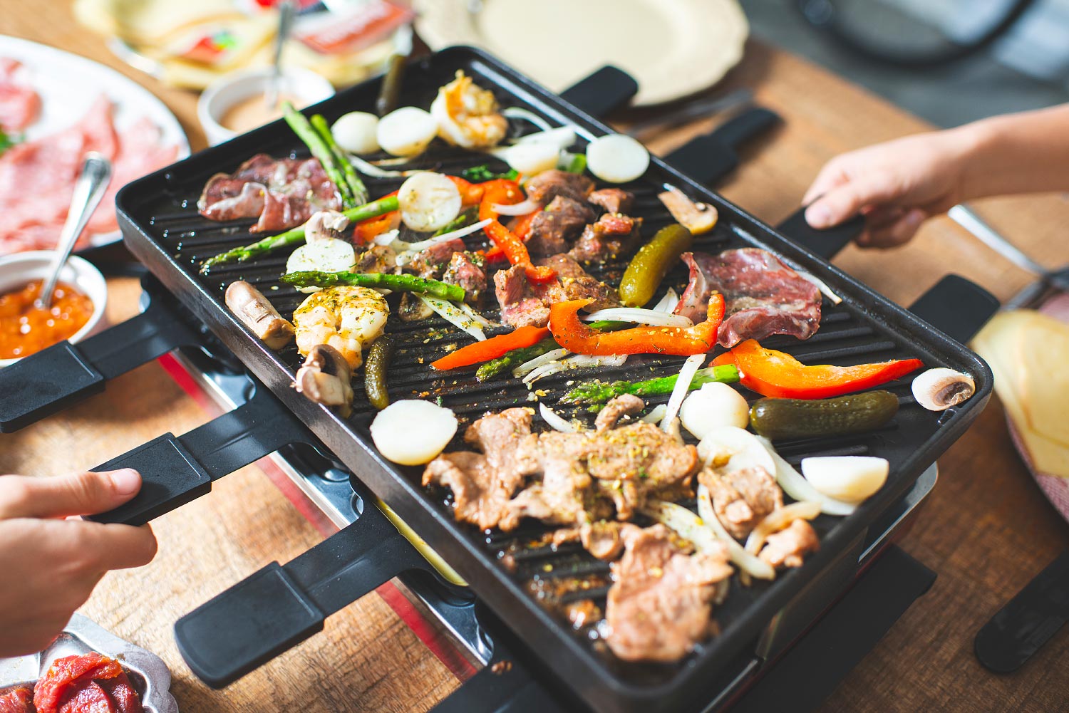 Des mains sur les poêlons autour d'une plaque de raclette bien garnie
