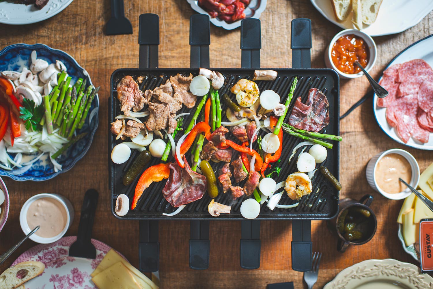 Table de raclette vue de haut : l'appareil au centre, la plaque garnie, les charcuteries, les légumes et les sauces